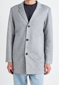 Uomo che indossa un blazer grigio chiaro con bottoni sopra una camicia nera e jeans azzurri chiari, in piedi davanti a uno sfondo bianco.