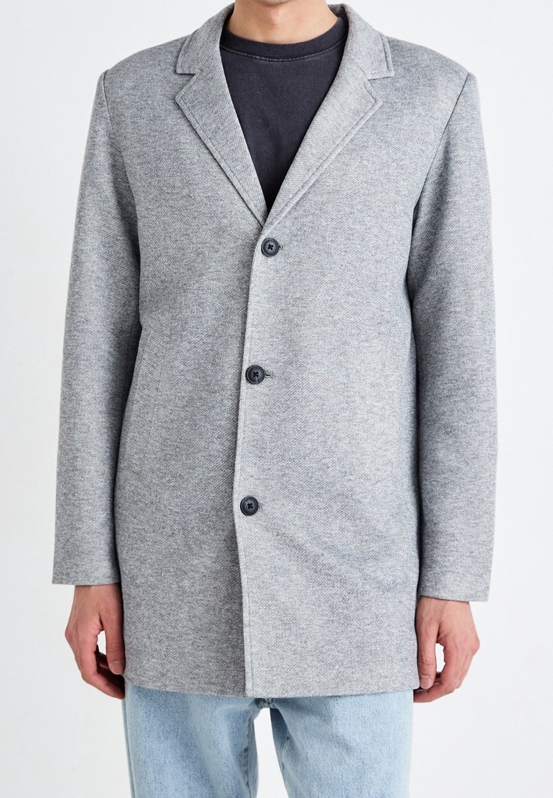 Uomo che indossa un blazer grigio chiaro con bottoni sopra una camicia nera e jeans azzurri chiari, in piedi davanti a uno sfondo bianco.