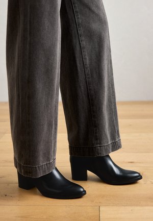 Zwarte leren enkellaarsjes met een puntige neus en blokhak, gecombineerd met grijze wijde broek, tegen een houten vloer.