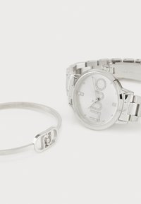 Zilverkleurig horloge met een ronde kast, een met kristallen bedekte wijzerplaat en een schakelarmband. Vergezeld van een zilveren manchetarmband met een logo-accent.