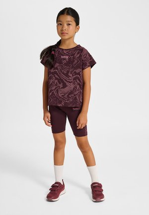 Ung pige står iført en vinrød mønstret T-shirt, matchende shorts, hvide sokker og vinrøde sneakers på en hvid baggrund.