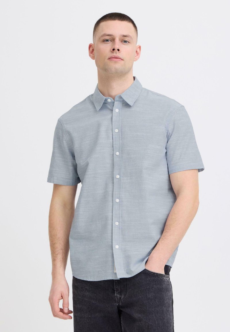 Άνδρας με κοντά μαλλιά που φοράει ανοιχτό μπλε πουκάμισο με κοντά μανίκια και μαύρο τζιν, στέκεται με το ένα χέρι στην τσέπη.