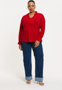 Rød bluse med lange ærmer, V-hals og flæsekant; parret med blå vide jeans med opsmøgte ben. Enkle hvide sandaler.