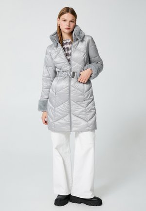 Cappotto isolato argento con vita punto vita, colletto in pelliccia morbida e motivo chevron testurizzato, abbinato a pantaloni bianchi a gamba larga e stivali neri.