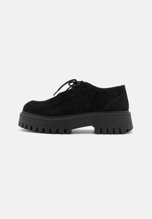 Scarpe nere in suede con lacci e suola a piattaforma caratterizzata da scanalature profonde, punta arrotondata e cuciture resistenti lungo il bordo.