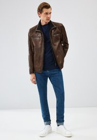 Veste en cuir marron avec des poches zippées, portée sur une chemise bleu marine. Assortie avec un jean bleu et des baskets blanches, affichant un look décontracté.