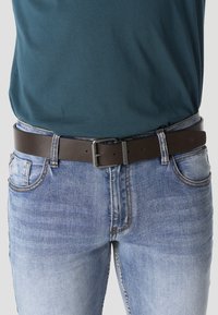 Persona con jeans azul claro y un cinturón de cuero marrón oscuro con hebilla metálica cuadrada, combinado con una camisa color verde azulado metida por dentro.