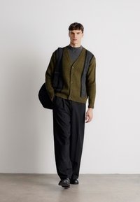 Καρδία με σχέδιο σε ελαιόλαδο και μαύρο, με κουμπιά μπροστά, φορεμένο πάνω από γκρί ζιβάγκο; συνδυασμένο με μαύρο παντελόνι φαρδιάς γραμμής και μαύρα παπούτσια.