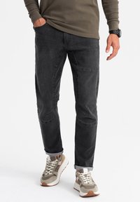 Pantalones de mezclilla negros con un corte slim, que cuentan con parches en las rodillas, puños casuales y bolsillos laterales, combinados con zapatillas deportivas de color claro.