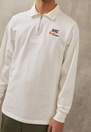 Homme portant un polo de sport Nike blanc à manches longues avec un col zippé et un logo Swoosh orange.