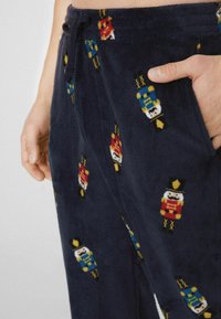 Granatowe polarowe spodnie do piżamy z wzorem dziadka do orzechów w kolorach czerwonym, niebieskim i żółtym. Posiadają boczne kieszenie oraz ściągacz w talii ze sznurkiem.