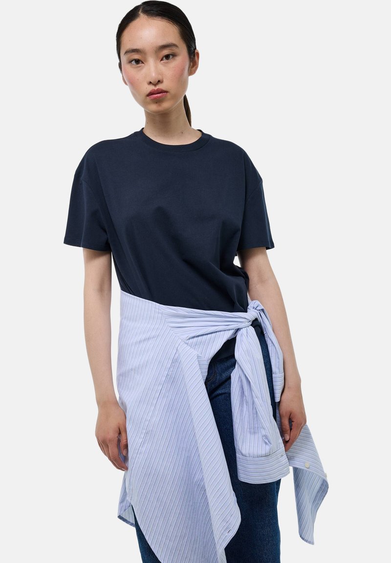 T-shirt bleu marine à manches courtes superposé sous un gilet rayé bleu clair et blanc avec un nœud à la taille, associé à un jean en denim sombre.