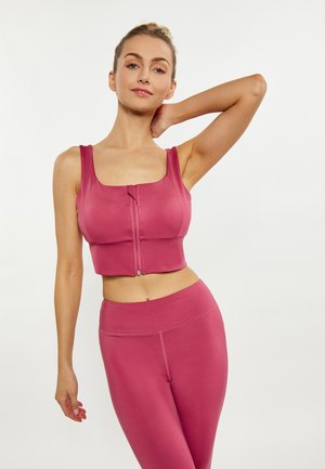 Femeie purtând crop top roz cu fermoar și colanți cu talie înaltă asortați, pozând cu o mână în spatele capului pe un fundal simplu.