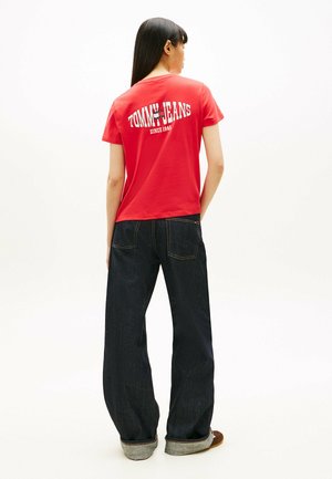 Donna con capelli lunghi e scuri che indossa una maglietta rossa Tommy Jeans e jeans denim scuri larghi, in piedi di spalle su uno sfondo bianco.