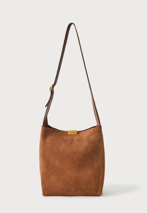 Rjava ramenska torba iz semiša z nastavljivim trakom in zlatim pravokotnim logotipom na vrhu na sredini, stoji ob enobarvni podlagi.