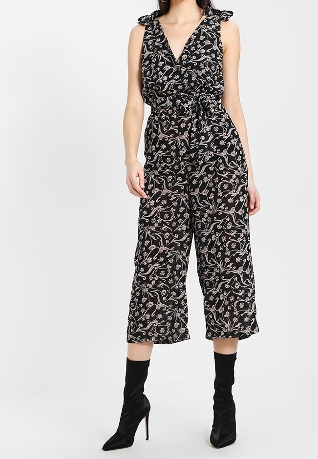 Combinaison noire à motif floral, avec bretelles volantées, taille cintrée et jambes larges raccourcies, associée à des bottes noires montantes jusqu'aux genoux.