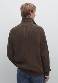 Brązowy sweter z prążkowanego dzianiny z wysokim kołnierzem, długimi rękawami i luźnym krojem, wykonany z fakturowanej tkaniny. Noszony z dżinsowymi spodniami.