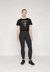 Juodas trumpas marškinėlis su auksiniu "GUESS" logotipu, derinamas su aukštu liemeniu juodomis leginsomis ir juodais sportiniais batais. Lygi medžiaga, prigludusi forma.