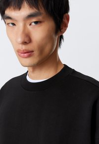Ung mand med kort sort hår iført en sort crewneck sweater over en hvid skjorte mod en hvid baggrund.