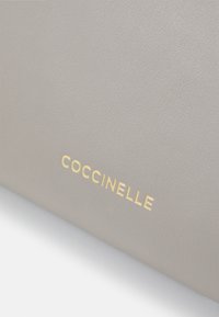 Grå læderhåndtaske med en struktureret overflade, forsynet med et guldfarvet præg-logo "COCCINELLE" på forsiden.