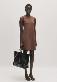 Bruine mouwloze jurk met een eenvoudig ontwerp, gecombineerd met een grote zwarte tote bag. De jurk heeft een ontspannen pasvorm en een zachte textuur.
