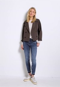 Brązowa podwójnie zapinana krótka kurtka, szary sweter, niebieskie dopasowane dżinsy i białe trampki z żółtymi podeszwami, stojąca na jednolitym tle.