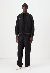 Black bomber jacket with gold accents, ribbed cuffs, worn over a black shirt. Paired with loose black cargo pants and white sneakers.