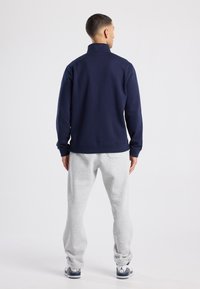 Navy pullover with a high collar, long sleeves, and ribbed hem; paired with light gray sweatpants and white athletic shoes.