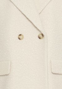 Manteau double boutonnage en crème, fabriqué en tissu texturé. Il dispose de deux grands boutons beiges et de deux poches avant avec des rabats.