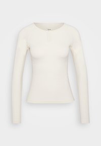 Haut ajusté à manches longues en tissu crème doux, avec un col rond et un design minimaliste. Présente un petit détail de fermoir à l'avant.
