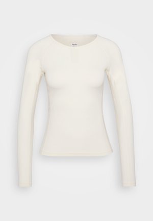 Haut ajusté à manches longues en tissu crème doux, avec un col rond et un design minimaliste. Présente un petit détail de fermoir à l'avant.