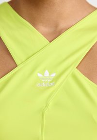 Neon žalios spalvos sportinis marškinėliai su kryžminiu nugaros dizainu, kuriuose yra baltas siuvinėtas Adidas logotipas ir lygus, tamprus audinys.