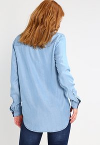 Ljusblå denimskjorta med långa ärmar, knäppkragar och en rundad nederkant. Bak- och framsidan visar en enkel design utan utsmyckningar.