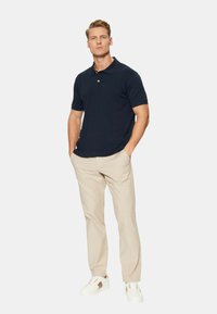 Polo shirt blu navy a maniche corte, pantaloni beige su misura e sneaker bianchi con dettagli marrone chiaro. Fit casual, texture morbida.