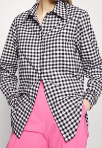 Chemise vichy noir et blanc avec col pointu, fermeture par boutons, manches longues et ourlet arrondi, associée à un pantalon rose.