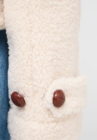 Lammfelljacke in Creme mit einer strukturierten, flauschigen Oberfläche. Verfügt über braune Knopfverschlüsse an den Bündchen, kombiniert mit Denim-Stoff.