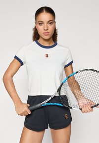 Λευκό cropped top με μπλε ραβδωτές λεπτομέρειες και ένα μικρό λογότυπο στο στήθος, συνδυασμένο με μαύρα σορτς και μια ρακέτα τένις.