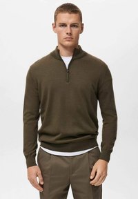 Suéter de color verde oliva con cremallera de un cuarto, hecho de material de punto suave, con un cuello alto y puños acanalados. Lleva encima una camisa blanca.
