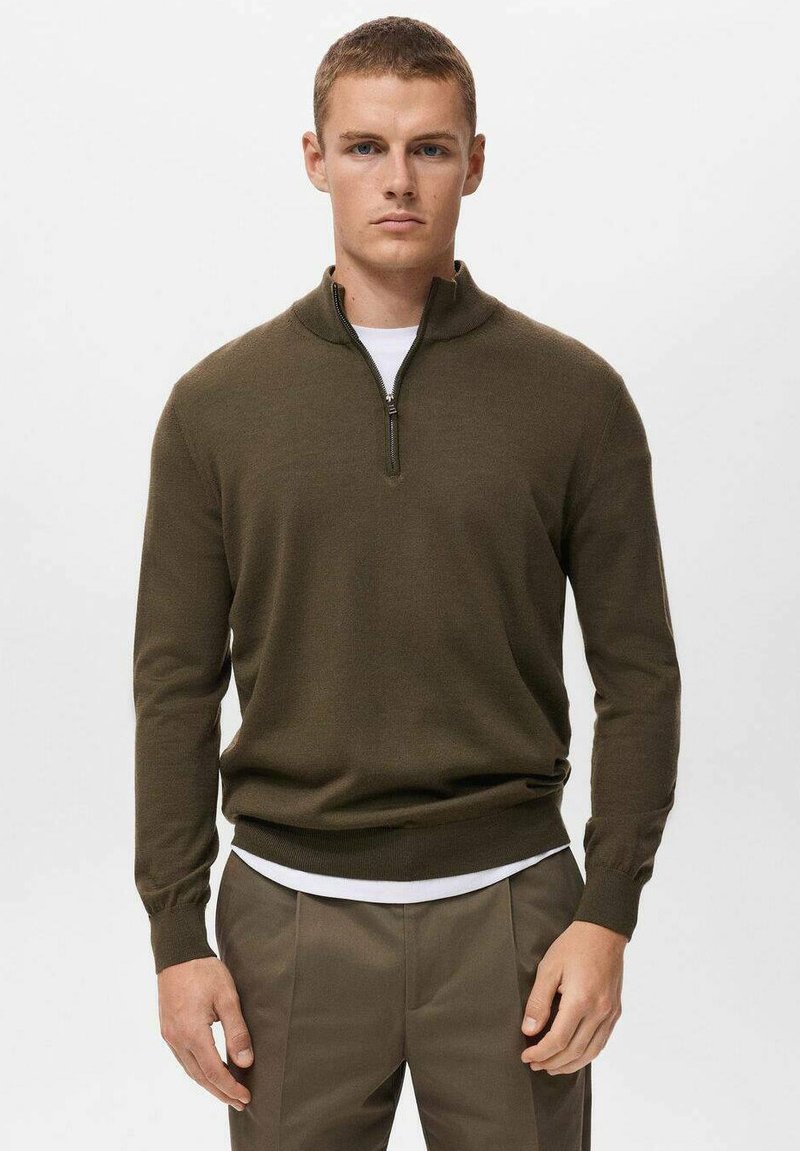 Suéter de color verde oliva con cremallera de un cuarto, hecho de material de punto suave, con un cuello alto y puños acanalados. Lleva encima una camisa blanca.