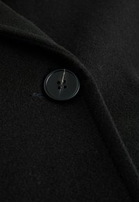 Fekete gyapjú kabát anyaga, amelyen közelről látható egy nagy fekete műanyag gomb négy lyukkal, mellette egy sima gallér él.