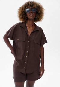 Czarnoskóra kobieta z kręconymi włosami nosi ciemnobrązową koszulę z guzikami i dopasowane szorty, uzupełnione dużymi okularami przeciwsłonecznymi i kolczykami w kształcie kół.