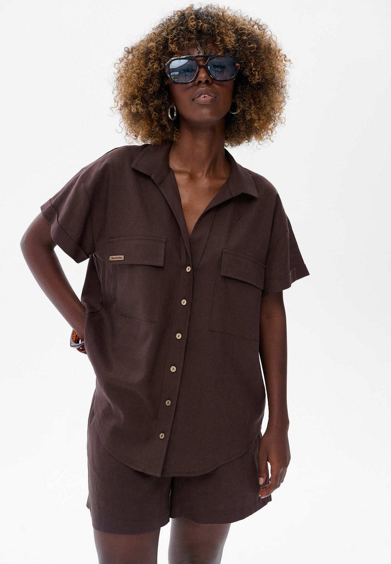 Czarnoskóra kobieta z kręconymi włosami nosi ciemnobrązową koszulę z guzikami i dopasowane szorty, uzupełnione dużymi okularami przeciwsłonecznymi i kolczykami w kształcie kół.