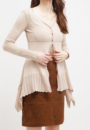 Vrouw draagt een beige vest met getande randen over een bruine suède rok, terwijl ze de knopen aan de voorkant vastmaakt.
