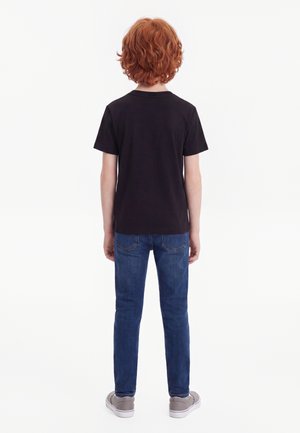 Barn med krøllet rødt hår iført en sort t-shirt, blå jeans og grå sneakers står med ryggen mod på en ensfarvet hvid baggrund.