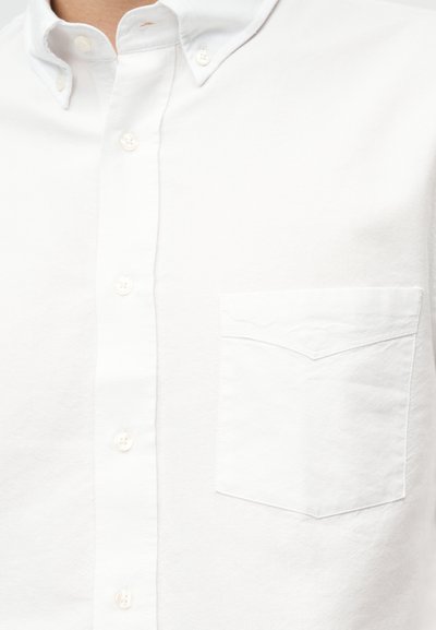 Κοντινή λήψη ενός λευκού πουκάμισου με κουμπιά, με κουμπωμένο γιακά και τσέπη στο στήθος στην αριστερή πλευρά.