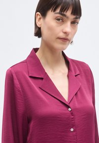 Blouse bordeaux à texture soyeuse, col classique et manches longues. Dotée de boutons sur le devant et d'une coupe légèrement décontractée.