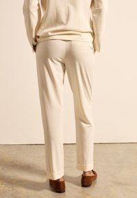 Cremefarbene maßgeschneiderte Hose mit geradem Bein, umgeschlagenem Saum und weicher Textur, kombiniert mit braunen Wildleder-Slippern. Neutraler Hintergrund.