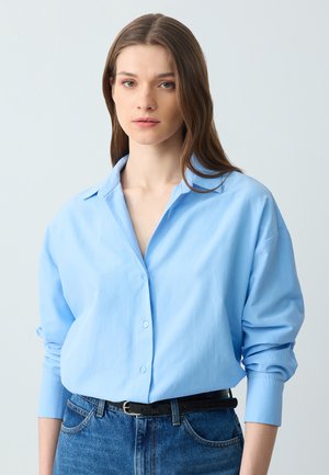 Lichtblauw overhemd met kraag, lange mouwen en een ontspannen pasvorm, gecombineerd met high-waisted blauwe jeans en een zwarte riem.