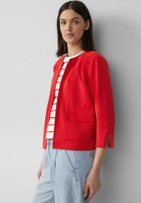 Rode jas met lange mouwen, ronde halslijn en voorkeurszak; gedragen over een witte en gestreepte shirt; textiele stof.