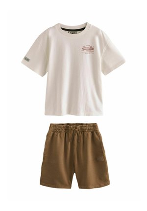 Niño joven con camiseta beige, pantalones cortos marrones y zapatillas blancas, de pie mientras ajusta una camisa de cuadros atada alrededor de su cintura sobre un fondo liso.